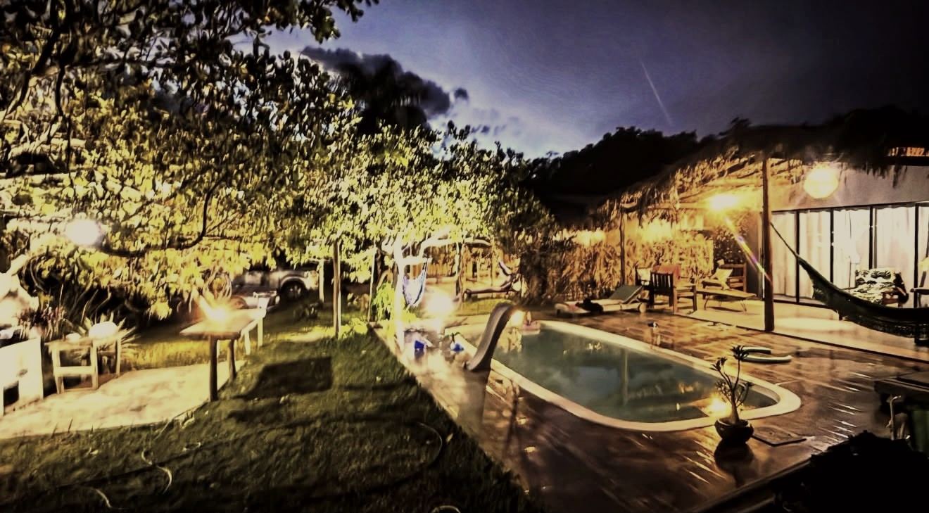 A pousada à noite, com a piscina iluminada, uma rede na varanda e as luzes acesas entre as árvores